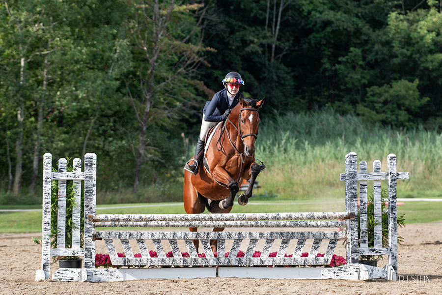 Girl with flowers on her helmet and red heart sunglasses riding a brown horse jumping