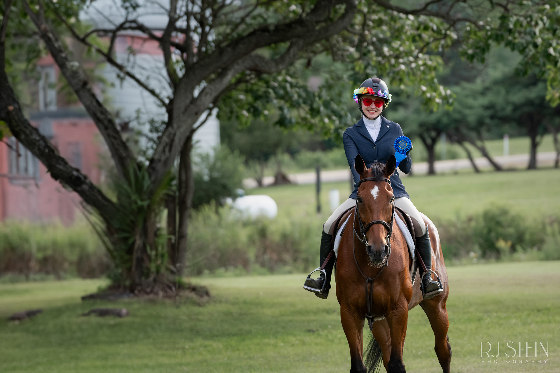 Girl with flowers on her helmet and red heart sunglasses smiling holding a blue ribbon while on top of a horse